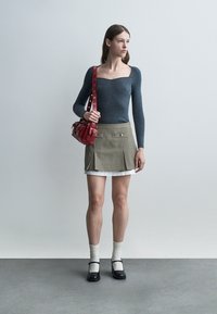 Mladá žena stojí opretá o jednoduchú stenu, oblečená v sivom tričku s dlhým rukávom, kockovanej mini sukni, bielych ponožkách, čiernych topánkach Mary Jane a drží červenú kabelku.