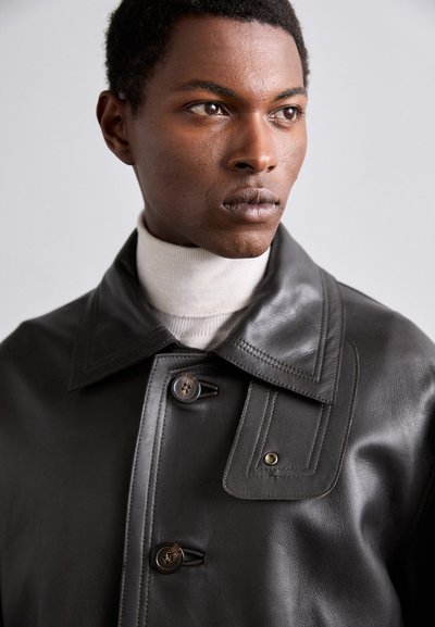 Sort læderfrakke med en bred krave, to knapper og en struktureret design. Båret over en lys beige turtleneck sweater.