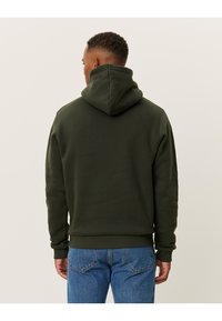 Mørkegrøn hættejakke med snøre i hætten, ribbede manchetter og talje, lavet af blødt materiale, parret med blå jeans.