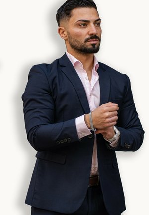 Dark navy suit jacket over a light pink shirt, features a pocket and silver bracelet. Man's hands adjust cuffs against a light backdrop.