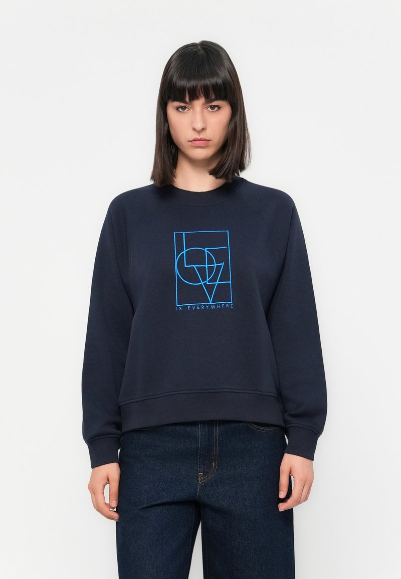 Sudadera de color navy con mangas raglán, que presenta un diseño gráfico azul y el texto "ESTÁ EN TODAS PARTES" en la parte frontal. Fabricada con una tela suave.