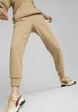 Beige sweatpants met een relaxte pasvorm, geribbelde manchetten, gemaakt van zacht materiaal, gecombineerd met beige en bordeauxrode sneakers, tegen een grijze achtergrond.