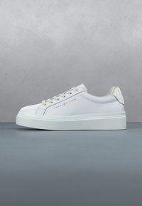 Baskets basses blanches avec lacets beiges, détail beige au talon, semelle épaisse et logo Tommy Hilfiger doré sur le côté sur fond gris.
