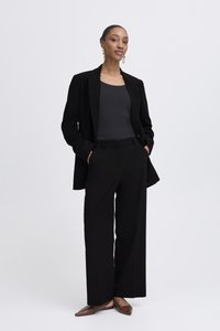 Črni krojeni suknjič čez črno majico brez rokavov, široke črne hlače in rjavi nizki čevlji. Preprost dizajn z gladkimi teksturami in minimalnimi poudarki.