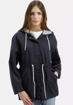 Młoda kobieta ubrana w granatową kurtkę z kapturem, białymi sznurkami, guzikami z przodu i bocznymi kieszeniami, stojąca na tle jasnego, jednolitego tła.