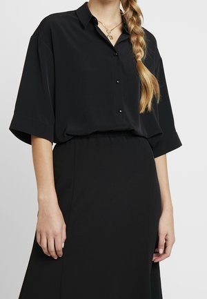 Woman wearing a loose black button-up shirt with elbow-length sleeves and a matching black skirt, plus a gold pendant necklace.