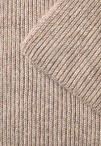 Beige ribbet strikket stof med en blød, tekstureret finish. Har skiftende tykke og tynde ribmønstre, der viser forskellige farvepletter.
