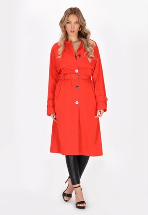 Rode trenchcoat met een ceintuur in de taille, grote knopen, lange mouwen en zijzakken. Contrast met zwarte leggings en hoge sandalen.