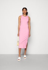 Vestido midi rosa sem mangas, ajustado com fenda lateral. Textura suave, tecido leve. Combinado com sandálias brancas de plataforma e uma bolsa amarela.