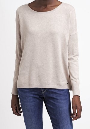 Kvinde iført en beige langærmet sweater med bådudskæring og blå jeans, stående mod en ensfarvet hvid baggrund.