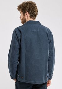 Veste en toile bleu marine avec un col, des poignets à boutons et une texture, dotée de poches pratiques et d'un design simple.