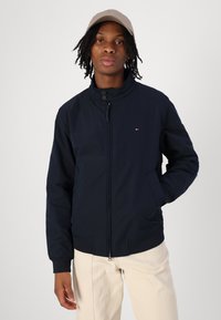 Veste bleu marine avec un col haut, fermeture éclair sur le devant et deux poches latérales ; poignets et ourlet élastiques. Portée avec un pantalon de couleur claire et une casquette.
