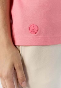 Camiseta de color rosa claro hecha de algodón, con un pequeño logo JOTT en relieve en el dobladillo inferior izquierdo y una textura suave.