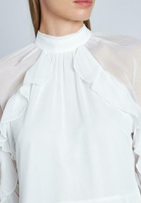 Camicia bianca con colletto alto, dettagli arricciati lungo lo scollo e le maniche, realizzata in tessuto leggero e testurizzato.