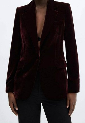 Persona che indossa un blazer in velluto bordeaux scuro sopra una maglia nera e pantaloni neri, in piedi davanti a uno sfondo chiaro e semplice.