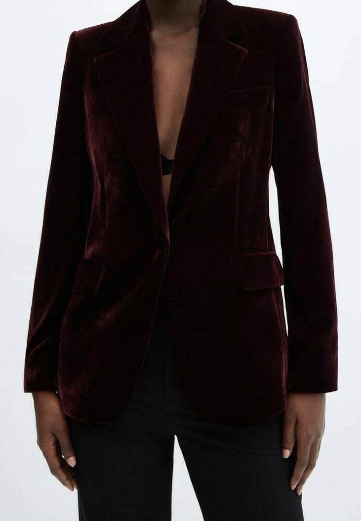 Personne portant un blazer en velours bordeaux foncé sur un top noir et un pantalon noir, debout devant un fond clair uni.