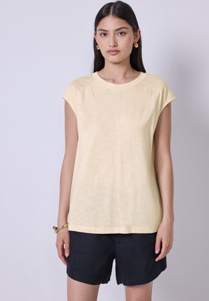 Top giallo chiaro a maniche corte in tessuto morbido, con scollo rotondo e una leggera texture. Indossato con pantaloni corti scuri, per un look casual.