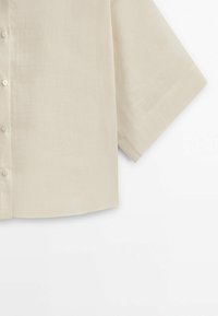 Beige liinapaita, jossa on lyhyet hihat, pyöristetty helma ja teksturoitu pinta. Paidassa on kuusi nappia etuhalkiolla.