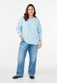 Hellblaues Strickpullover mit einem Diamantmuster, kombiniert mit lockeren blauen Jeans und schwarzen Slippern mit dekorativen Akzenten.