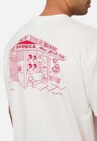 Weißes Baumwoll-T-Shirt mit einer roten Lin Zeichnung eines Brooklyn Deli-Markts, das einen Ladenfront mit Stühlen, Pflanzen und Kartons zeigt.
