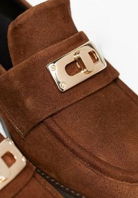 Mocassini in suede marrone con una trama spessa e dettagli in metallo dorato. La texture è morbida, con cuciture lungo i bordi.
