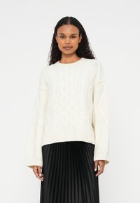 Jersey blanco de punto con patrones de trenzas y hombros caídos, combinado con una falda negra con pliegues. El jersey cuenta con dobladillos y puños acanalados.