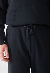Hanorac din bumbac negru, cu o buzunar mare frontal, cordon elastic la talie și o croială lejeră. Textură moale, cu detalii hardware minime.