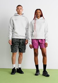 To personer står side om side. Personen til venstre har en lysegrå hættetrøje og mørke cargo shorts på. Personen til højre har en grå hættetrøje og pink leopardmønstret shorts på. Begge har sorte sneakers på.