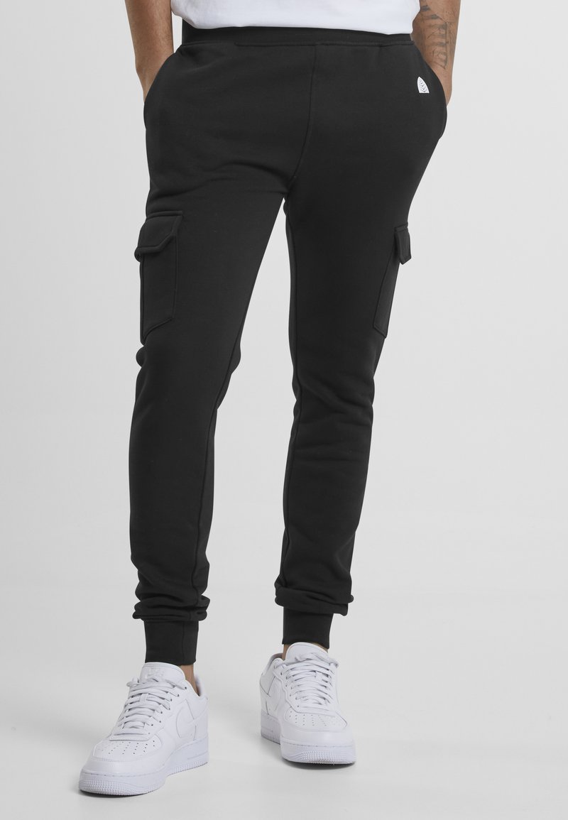 Black cargo joggers with tapered legs, featuring two side pockets. Worn with white sneakers and a plain white shirt. Smooth, stretchy fabric.