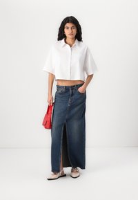 Camisa branca de botão cropped, saia de ganga de cintura alta com abertura frontal, mala vermelha e sapatos planos bege com detalhes pretos.