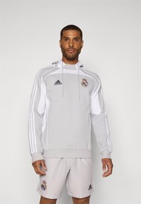 Siva Adidas dukserica s bijelim detaljima, logotip Real Madrida i kapuljača s vezicom. Par svijetlosivih kratkih hlača s logotipom.