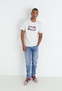 Witte korte- mouwen t-shirt met een Union Jack ontwerp op de borst, gecombineerd met blauwe spijkerbroeken en rood-witte sneakers.