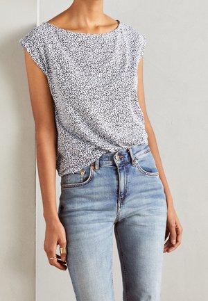 Femme portant un haut blanc sans manches avec des taches noires, rentré dans un jean bleu clair, debout contre un mur clair uni.