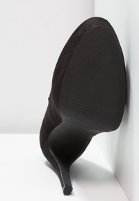 Chaussure à talon aigu en suede noir, avec un bout pointu et un talon stiletto élégant. La semelle est texturée avec des rainures pour une meilleure adhérence.