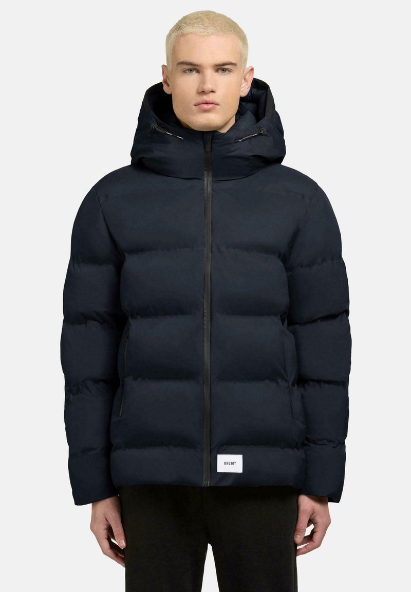 Giubbotto imbottito blu navy con cappuccio, dotato di chiusura frontale a zip e due tasche laterali. Texture morbida e trapuntata con etichetta del marchio sull'orlo.