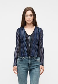 Γυναίκα με ίσια καστανά μαλλιά φοράει μπλε και μαύρη μπλούζα με μακριά μανίκια και σχέδιο, με κορδέλα στο μπροστινό μέρος, συνδυασμένη με ανοιχτό μπλε τζιν παντελόνι.