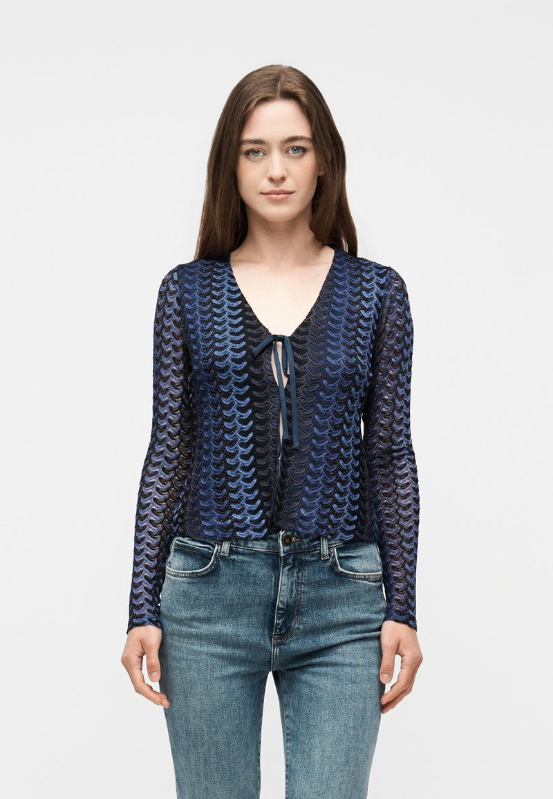 Γυναίκα με ίσια καστανά μαλλιά φοράει μπλε και μαύρη μπλούζα με μακριά μανίκια και σχέδιο, με κορδέλα στο μπροστινό μέρος, συνδυασμένη με ανοιχτό μπλε τζιν παντελόνι.