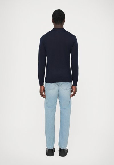 Uomo in piedi di spalle, che indossa una camicia nera a maniche lunghe, jeans azzurri chiari e scarpe nere, su uno sfondo bianco semplice.