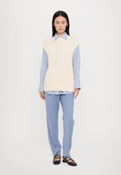 Kremasti pleteni bezrukavac s otvorenom mrežom, složen preko svijetloplavog prugastog košulje, u kombinaciji sa svijetloplavim hlačama i crnim balerinama.