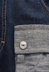 Εγγύς λήψη υφάσματος σκουρομπλε τζιν με γκρι θήκη με καπάκι, στερεωμένη με μεταλλικό κουμπί που φέρει χαραγμένο κείμενο.