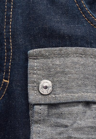 Εγγύς λήψη υφάσματος σκουρομπλε τζιν με γκρι θήκη με καπάκι, στερεωμένη με μεταλλικό κουμπί που φέρει χαραγμένο κείμενο.