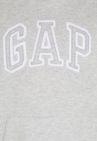Sudadera gris con las letras bordadas "GAP" en blanco y con un contorno texturizado. La tela parece suave con un acabado liso.