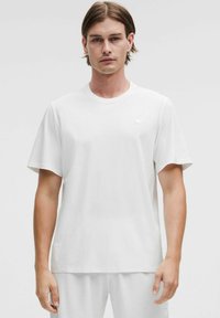 Camiseta blanca de manga corta con cuello redondo, que presenta una textura sutil. Detalle de logo pequeño en el lado izquierdo del pecho. Tejido ligero.