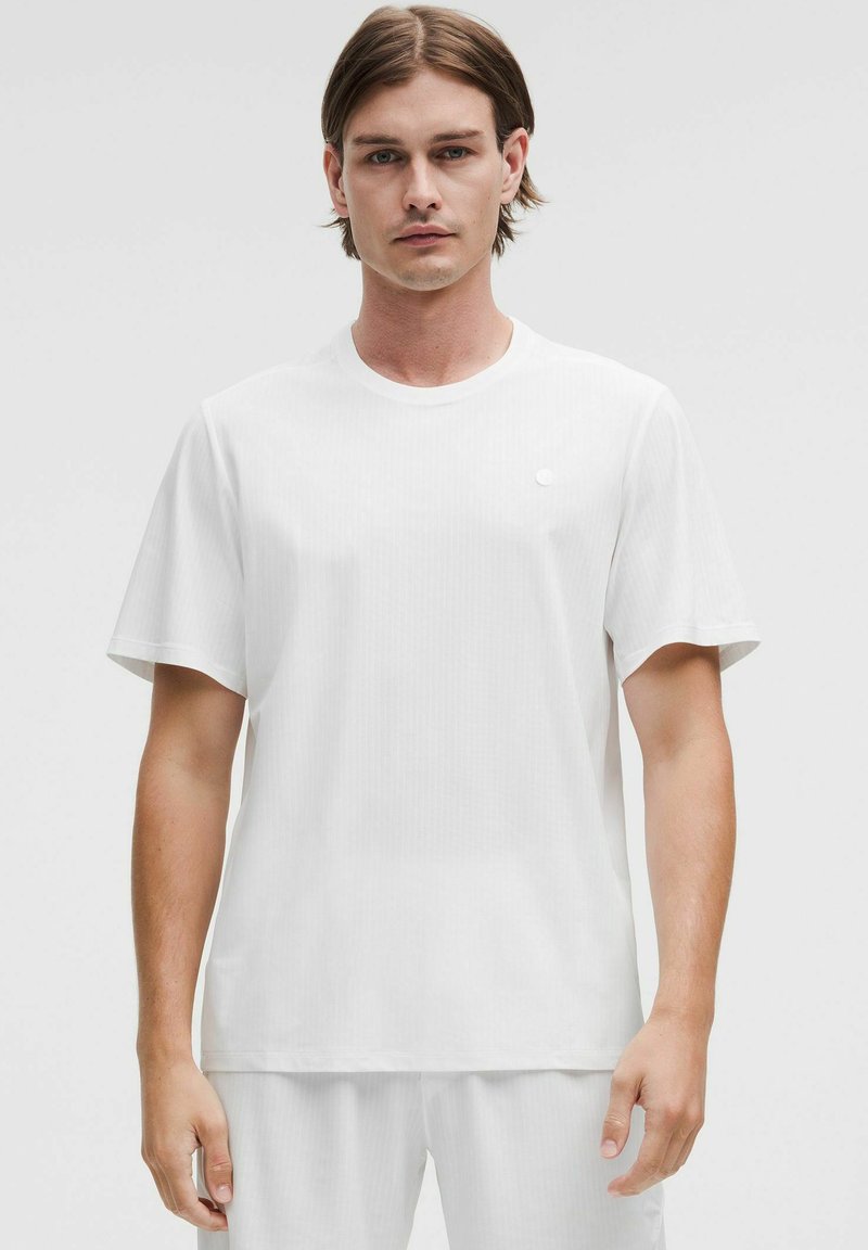 Camiseta blanca de manga corta con cuello redondo, que presenta una textura sutil. Detalle de logo pequeño en el lado izquierdo del pecho. Tejido ligero.