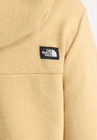 Veste en tissu côtelé beige avec un patch rectangulaire noir portant le logo blanc "The North Face" sur la poitrine.