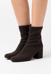 Bruna ankelstövlar gjorda av ett mjukt, sträckt material med en fyrkantig tå och en kraftig klack. Skrynklig design vid vristen för detalj.