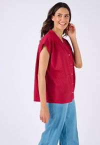 Top rosso a maniche corte con scollatura a V e chiusura con bottoni. Realizzato in cotone, presenta una vestibilità ampia e dettagli arricciati sulle spalle.