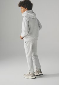 Chłopiec z kręconymi włosami, ubrany w jasnoszary hoodie, dopasowane dresy i białe sneakersy, stojący z boku na jednolitym tle.