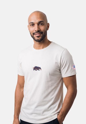 Uomo con barba corta e testa rasata che indossa una maglietta bianca con una piccola grafica dell'orso e una toppa sulla manica sinistra, in piedi davanti a uno sfondo bianco.