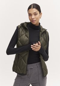 Gilet matelassé vert olive avec capuche, comportant une fermeture éclair sur le devant et des poches latérales, porté sur un haut à manches longues noir et un pantalon gris.
